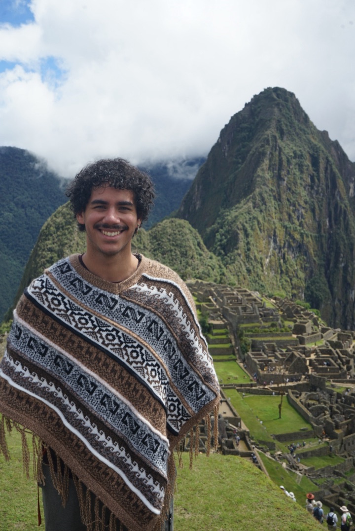 Josh Fonseca Rivera smiling in a woven poncho in front of Machu Picchu